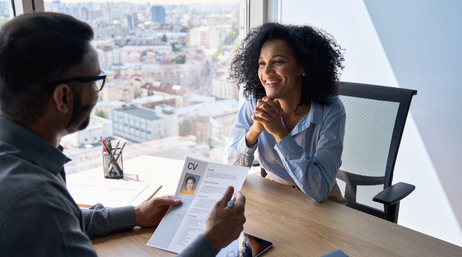 A woman sitting in a chair, being interviewed for a job. A man is holding up a copy of her CV.