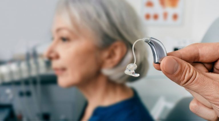 A hand holding up a single hearing aid. In the background, an older woman with shoulder-length grey hair is sitting on a chair.