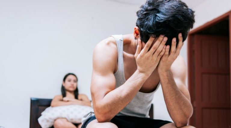 A man sitting on the end of a bed with his hand in his hands, looking frustrated. A woman is sitting in the background.